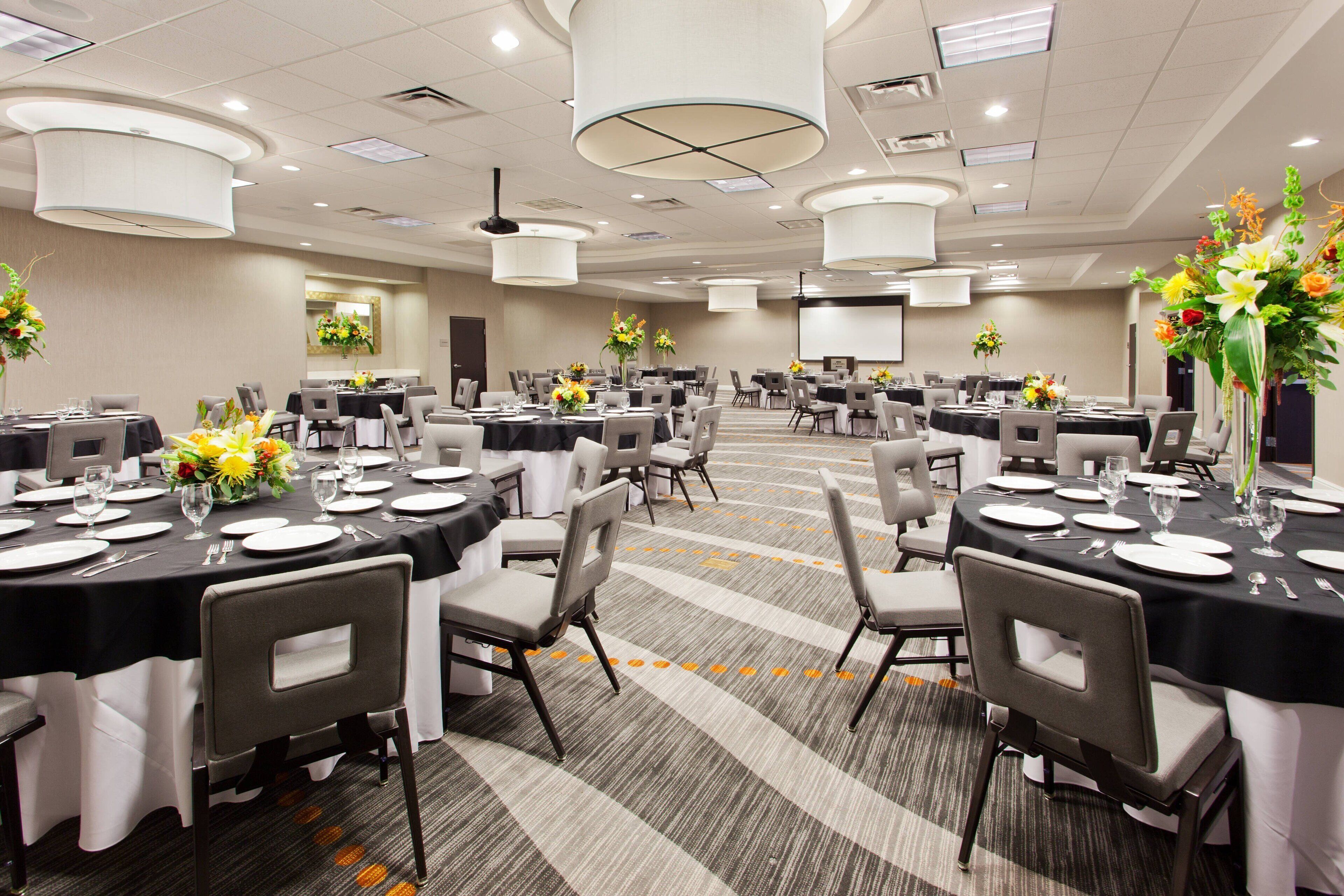 A conference room with multiple tables set for a meeting, equipped with chairs, tables, and floral centerpieces.