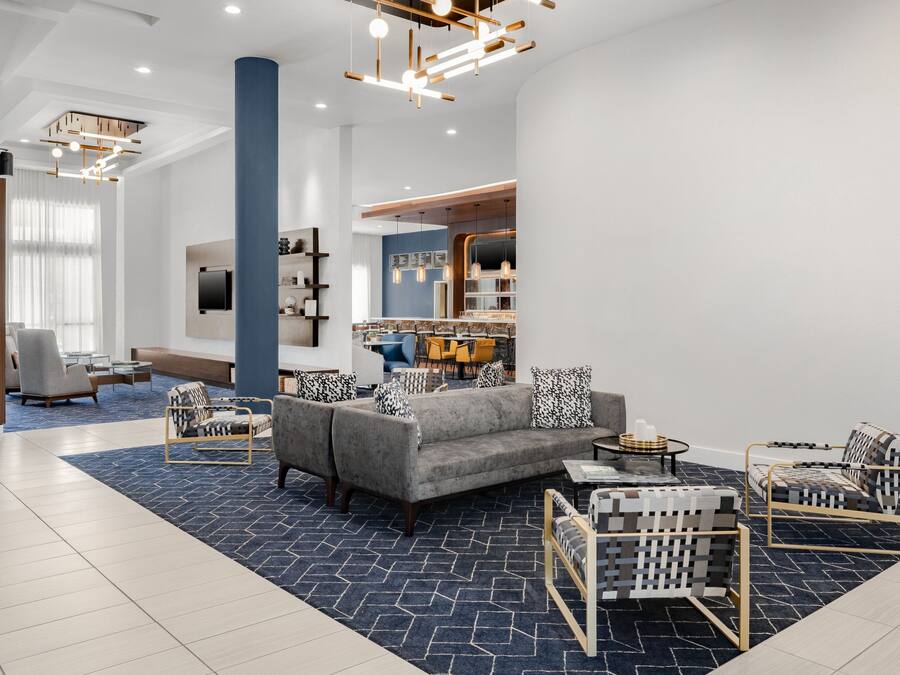 A modern hotel lobby with a grey sofa, patterned armchairs, a dining area, and a large pillar.