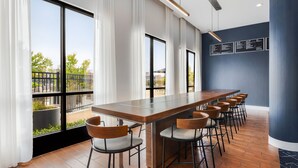 Zona de bar moderna con una larga barra de madera y varios taburetes, ventanas amplias con cortinas y una pared azul oscuro con paneles de menú.