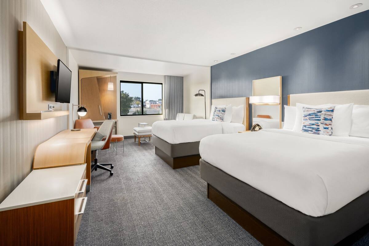 A hotel room with two beds, a desk, a chair, a TV, and a window with a view of buildings.