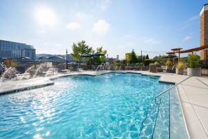 Una piscina con una fuente, rodeada de sillones de descanso y sombrillas, con un paisaje urbano al fondo.