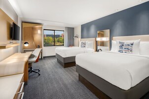 Habitación de hotel con dos camas, un escritorio, un televisor y un ventanal con vistas a árboles.