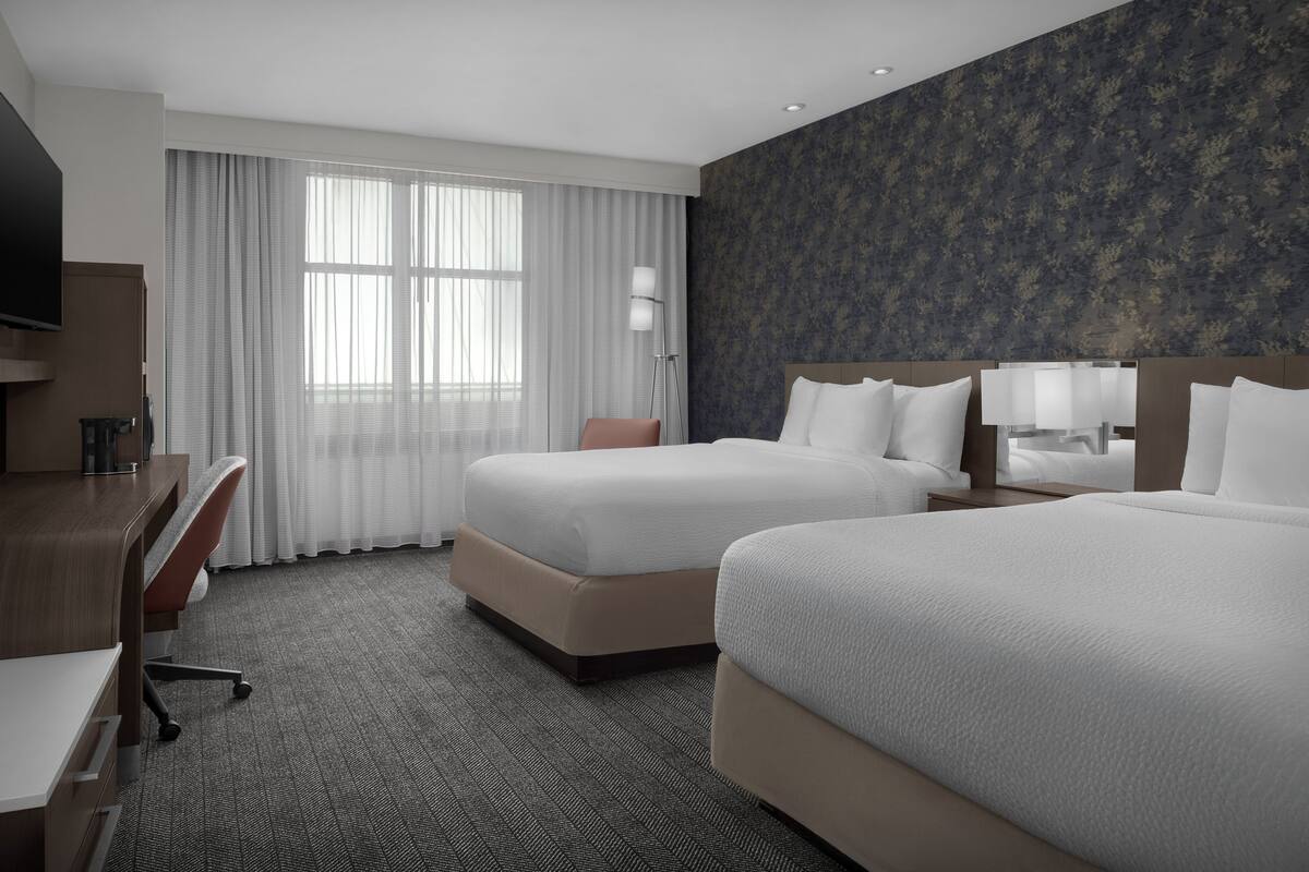A hotel room with two beds, a desk, a chair, a TV, and a large window with curtains.