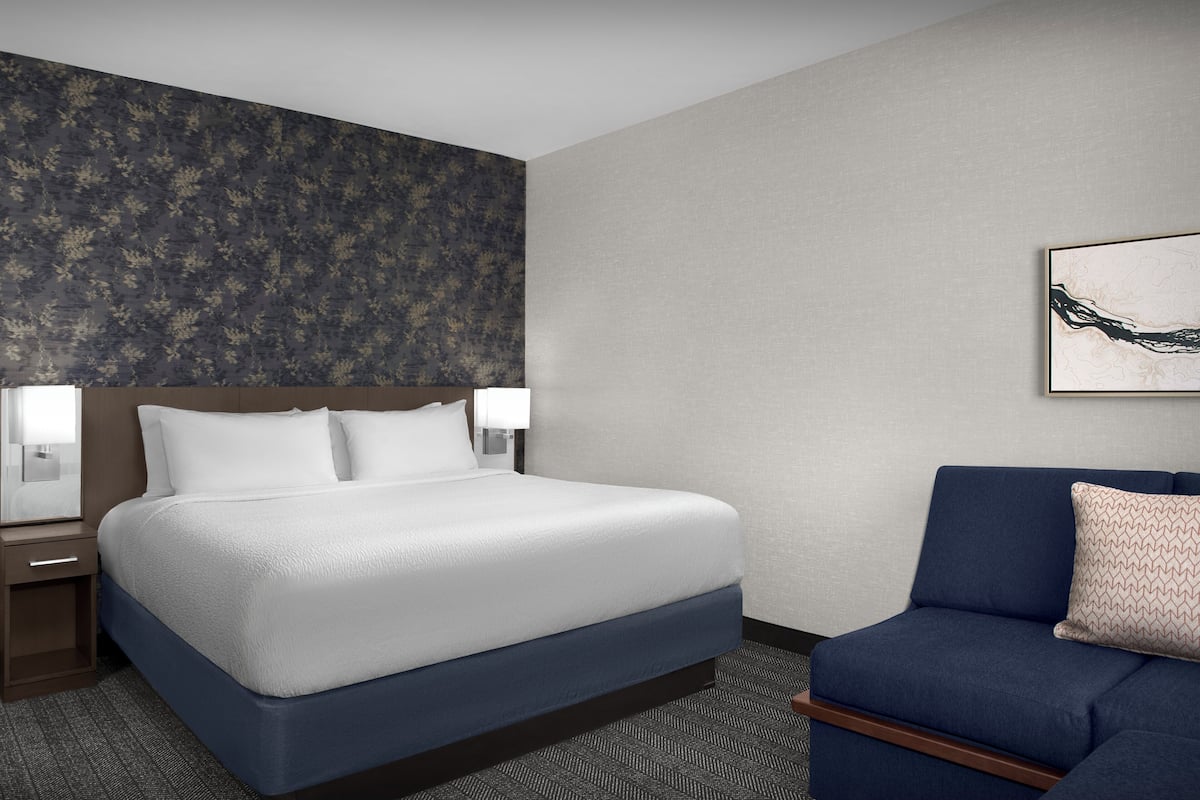 A hotel room with a large bed, a nightstand, a sofa, and a wall with a floral pattern.
