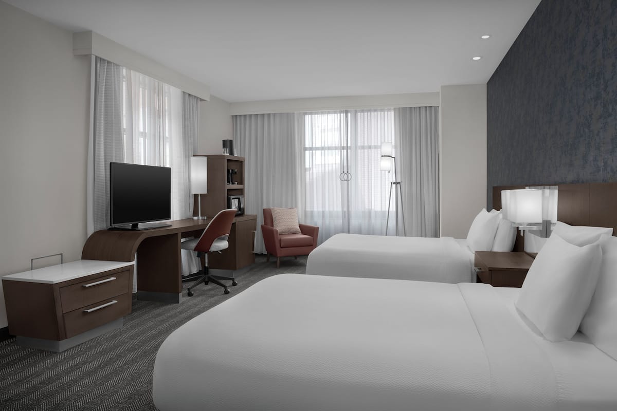 A hotel room with a large bed, a desk with a TV, a chair, a nightstand, and a lamp.