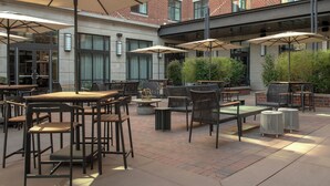 Een terras buiten met tafels en stoelen, parasols en een bakstenen gebouw op de achtergrond.
