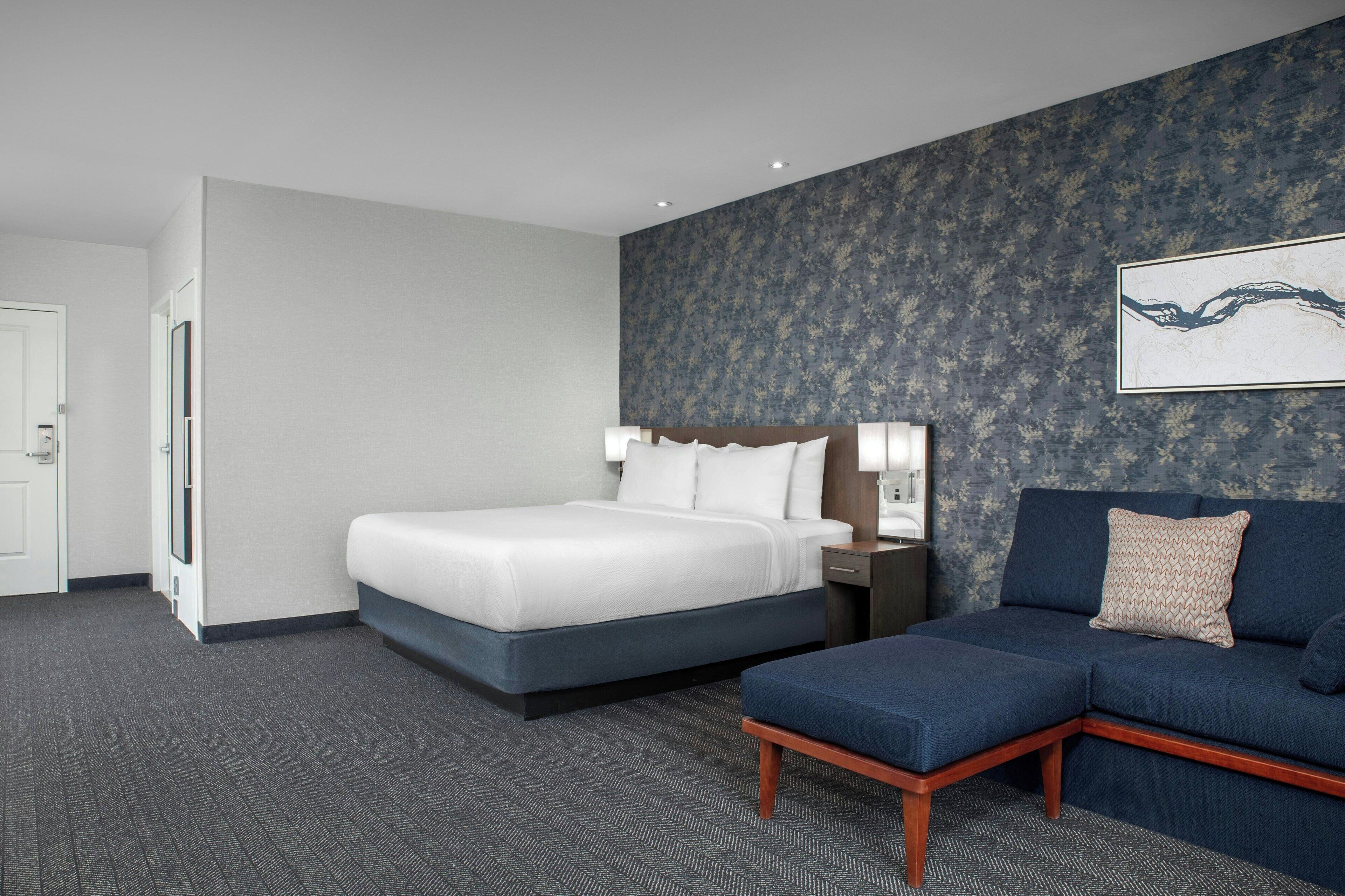 A hotel room with a bed, a sofa, a nightstand, and a wall-mounted lamp.