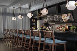 A bar area with multiple wooden bar stools, hanging pendant lights, and a well-stocked bar counter. - Courtyard by Marriott Richmond Downtown (Richmond)