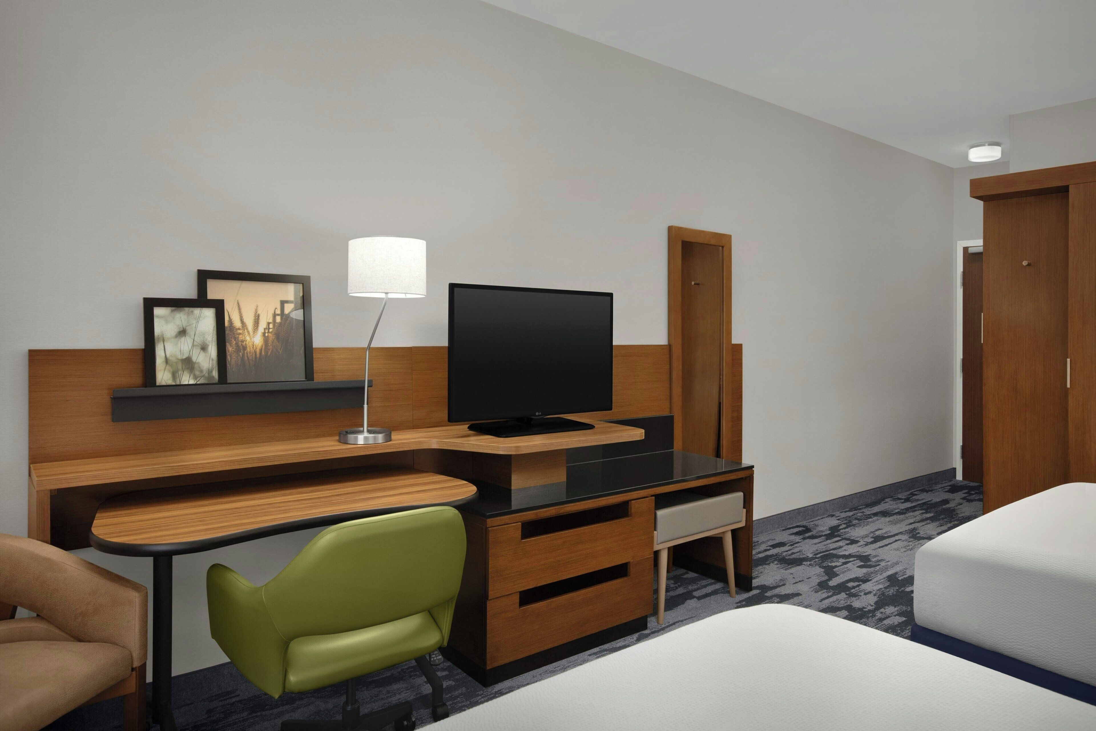 A hotel room with a desk, a television, a green chair, a bed, and a wardrobe.