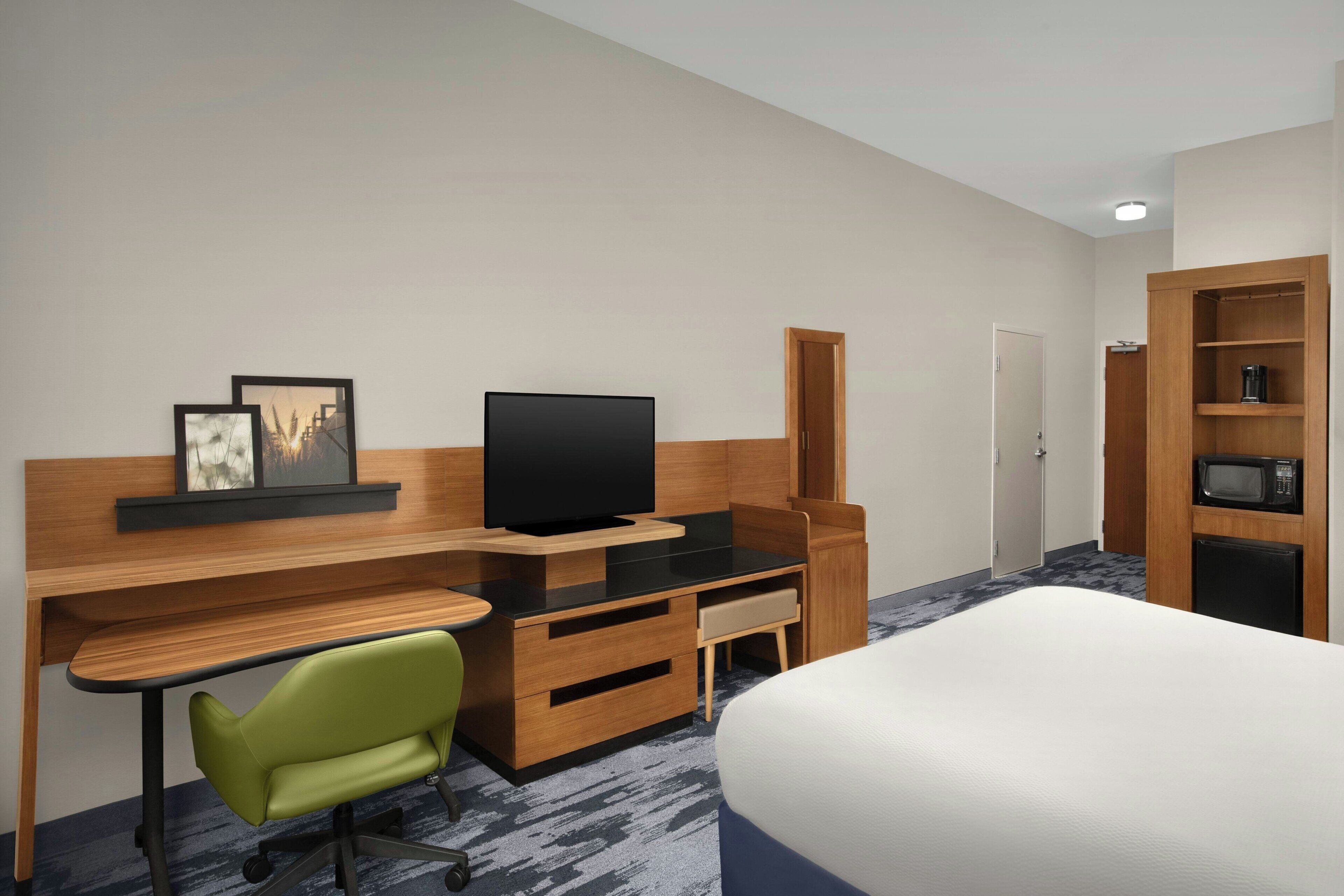 Habitación de hotel con cama, escritorio con televisor, una silla verde, armario y microondas.