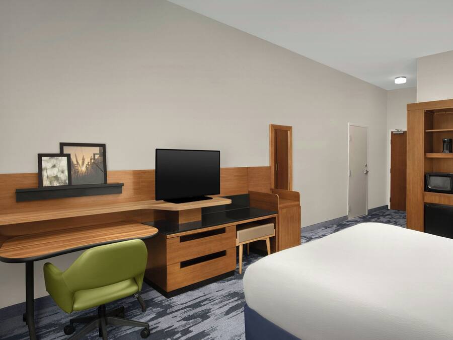 Habitación de hotel con cama, escritorio con televisor, una silla verde, armario y microondas.
