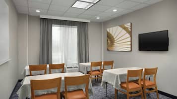 A conference room with tables and chairs, a large window with curtains, a flat-screen TV mounted on the wall, and a patterned carpet.