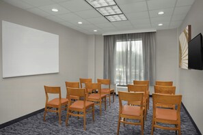 Une salle de conférence équipée d’un tableau blanc, d’un téléviseur à écran plat et d’une rangée de chaises en bois avec des coussins orange.