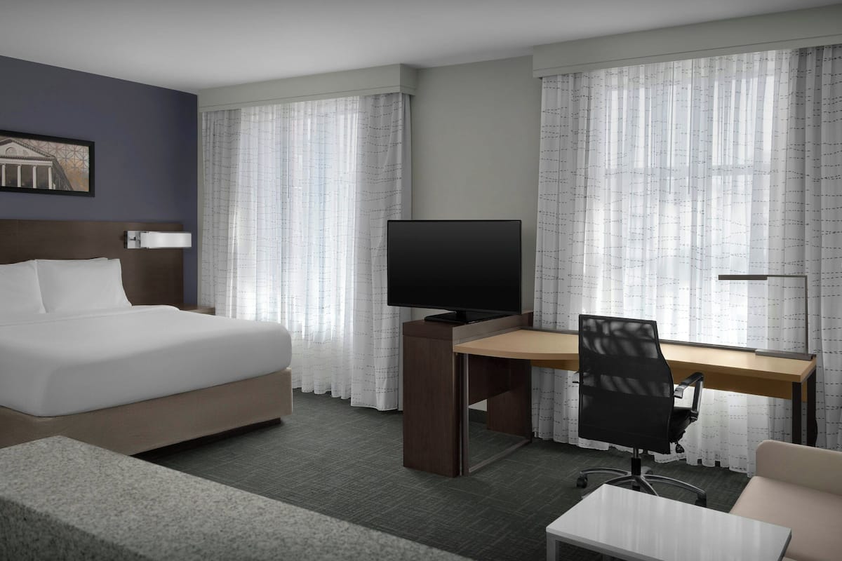 A hotel room with a bed, a desk with a TV, a chair, and a small table.