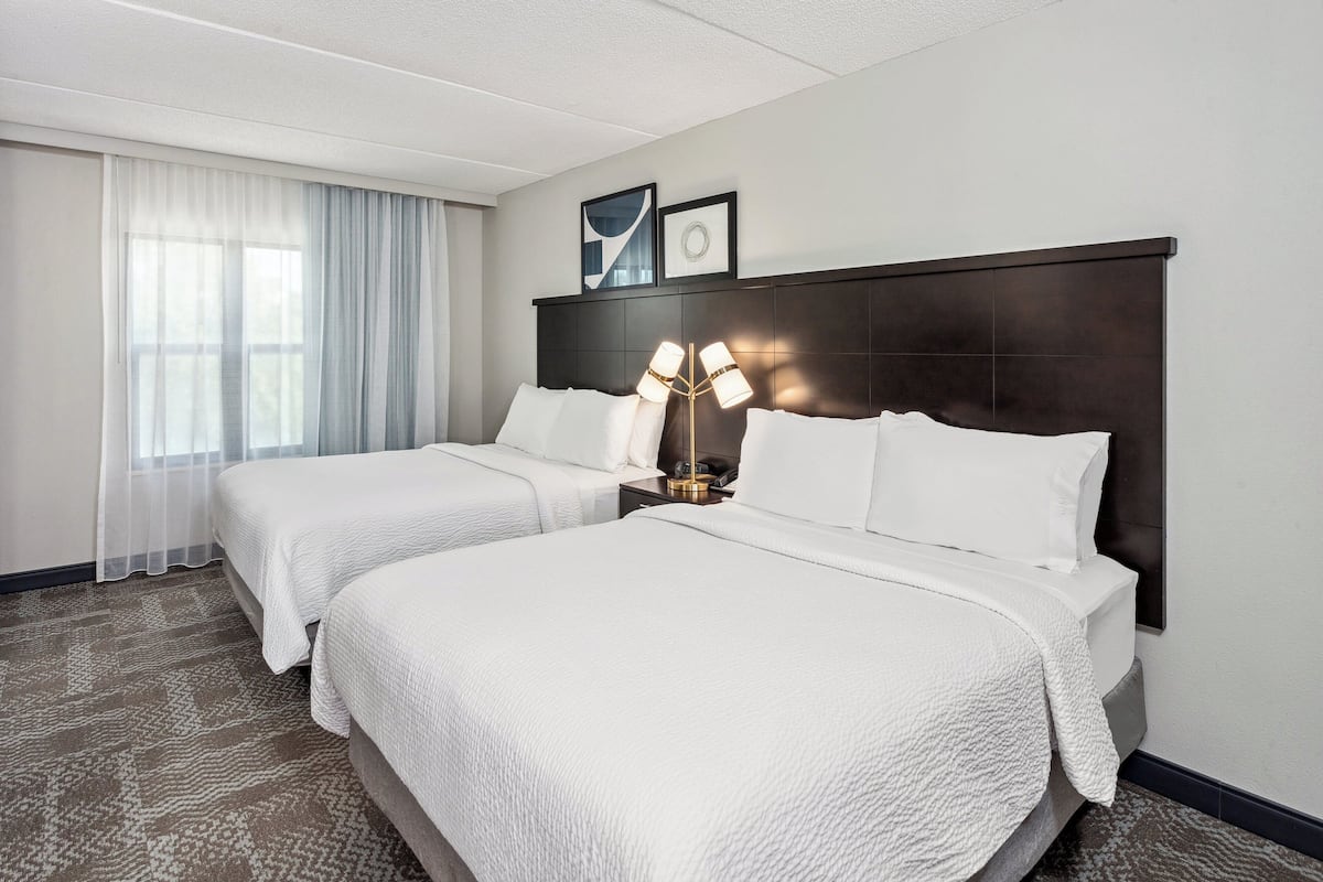 Une chambre d’hôtel avec deux lits, une grande fenêtre avec des rideaux légers, une tête de lit sombre et une lampe sur pied.