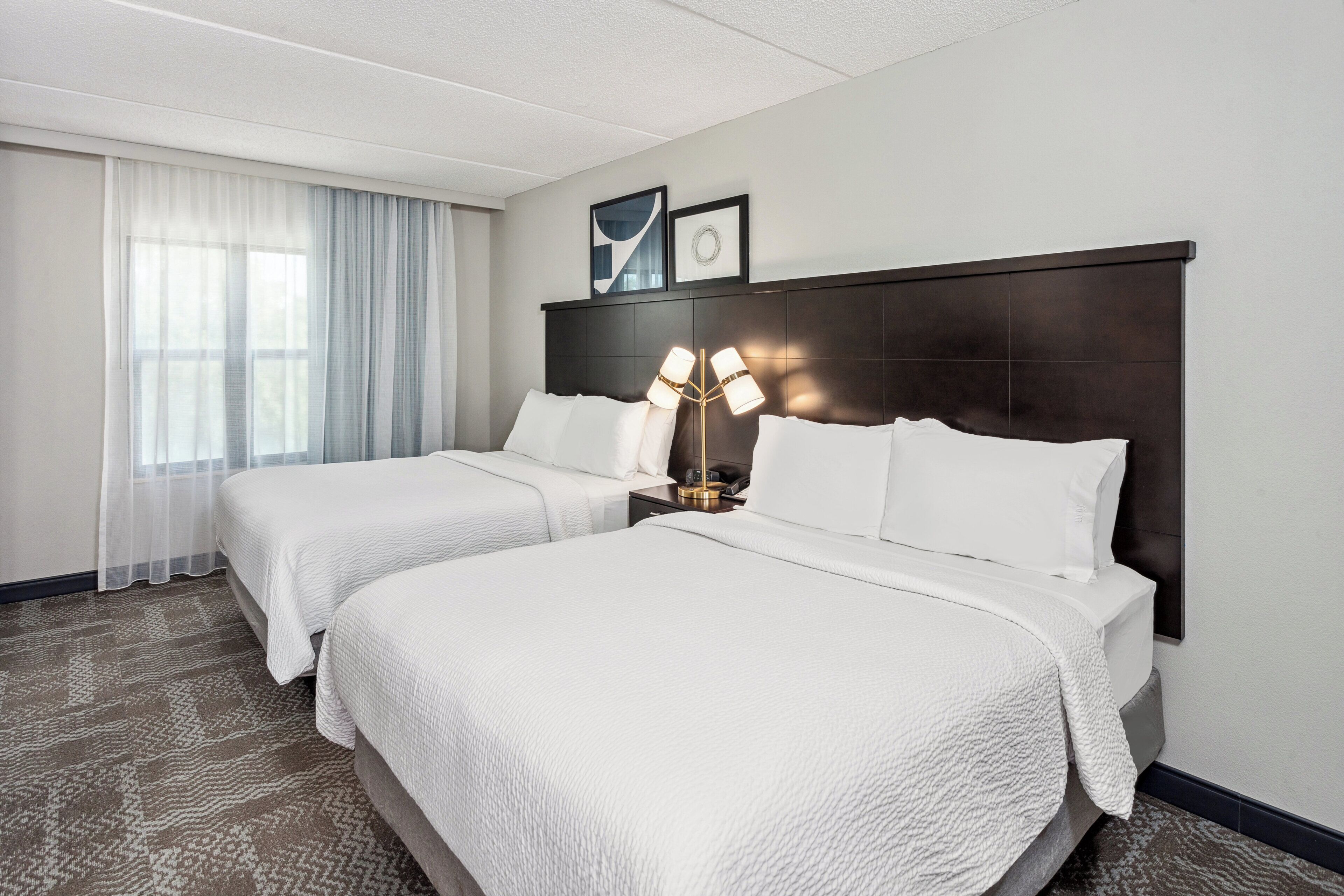 A hotel room with two beds, a large window with sheer curtains, a dark headboard, and a floor lamp.