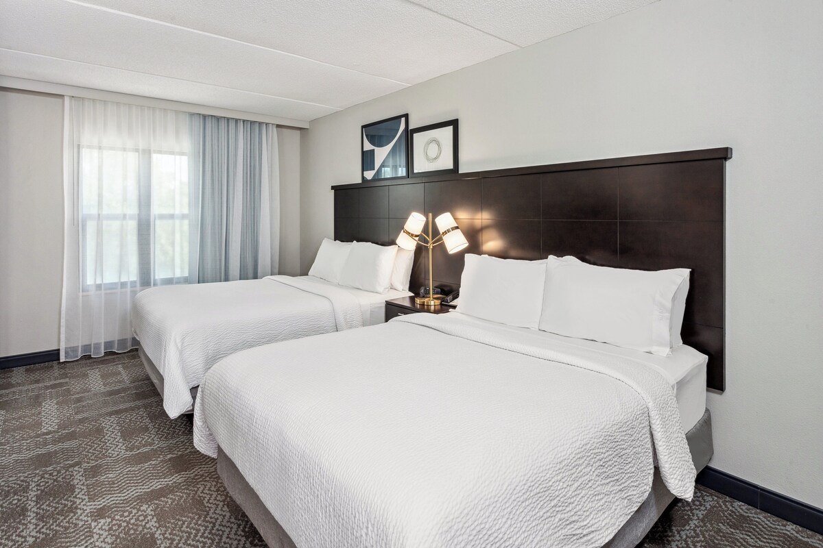 Une chambre d’hôtel avec deux lits, une grande fenêtre avec des rideaux légers, une tête de lit sombre et une lampe sur pied.