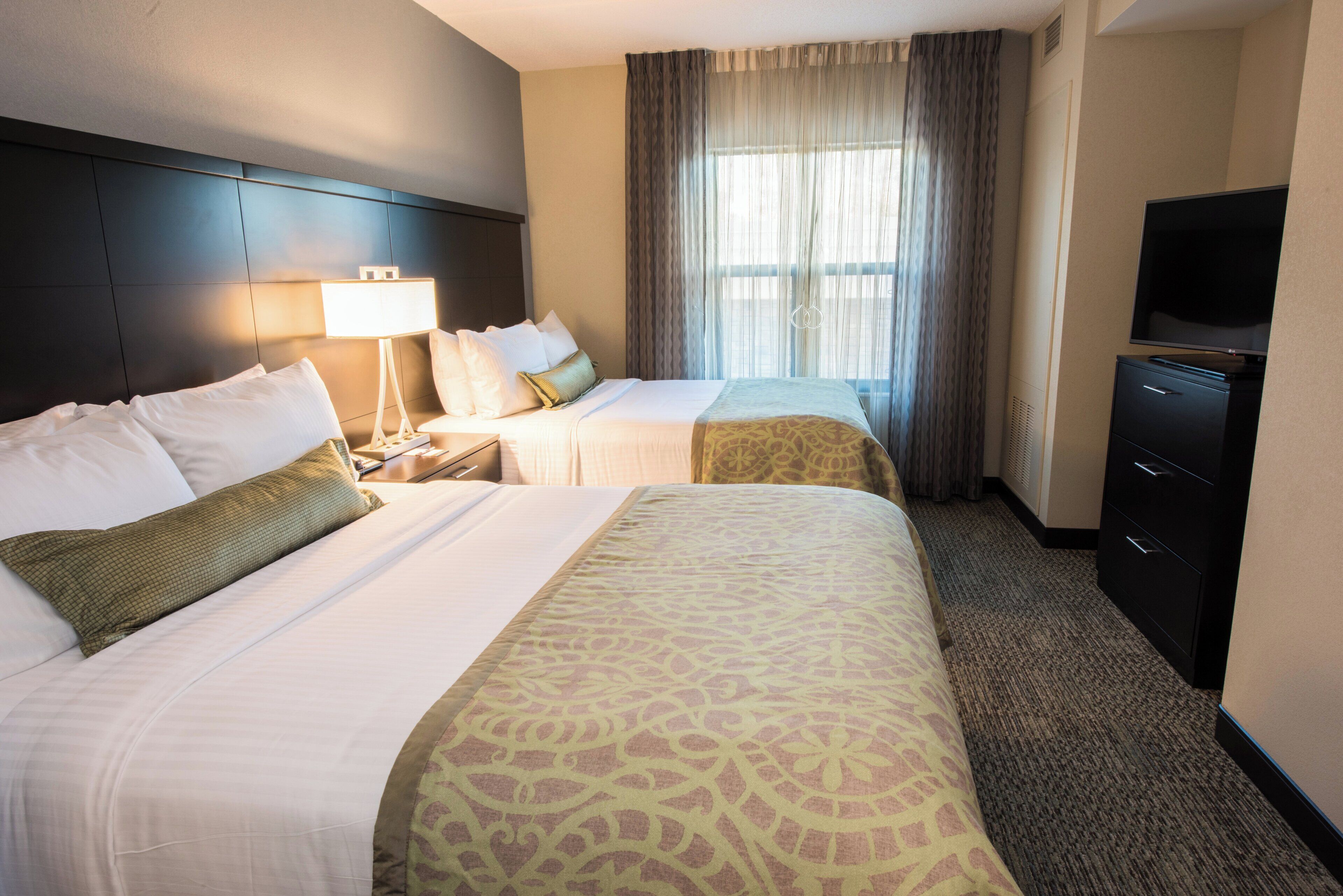 A hotel room with two beds, a TV, a desk, and a window with curtains.