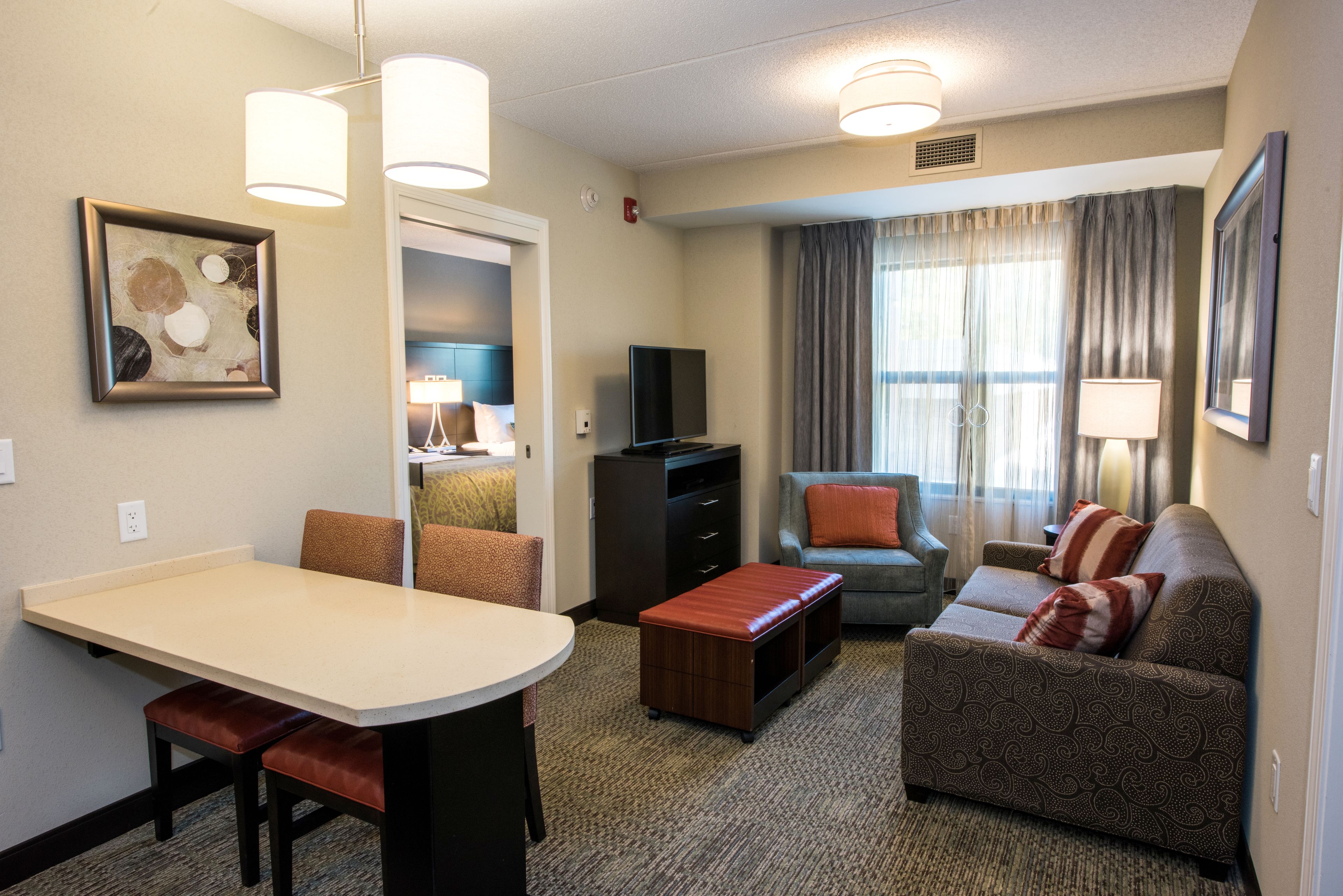 Une chambre d’hôtel comprenant un coin repas, un ensemble de canapés, une télévision et une grande fenêtre avec des rideaux.