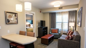Hotelkamer met een eethoek, zithoek, televisie en een groot raam met gordijnen.