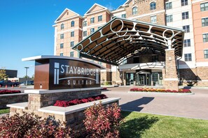 Entrada de un hotel con un cartel de Staybridge Suites, un paseo techado y macizos de flores ajardinadas.