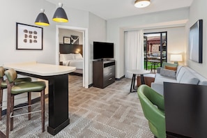 Habitación de hotel moderna con comedor, sofá, televisor y balcón con mobiliario exterior.