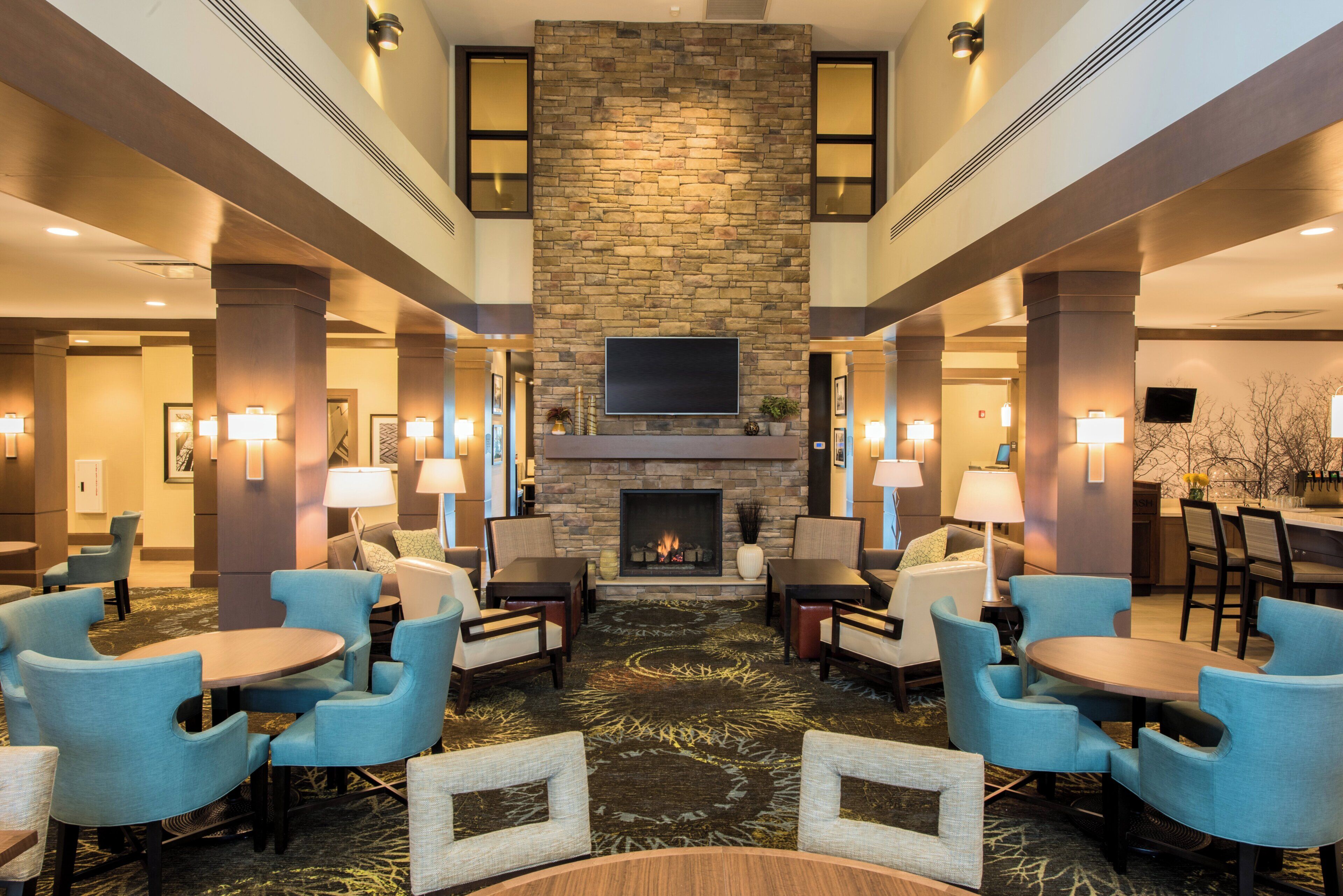 A hotel lobby with a stone fireplace, multiple seating areas with chairs and tables, and a flat-screen TV mounted above the fireplace.
