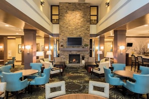 Un vestíbulo de hotel con chimenea de piedra, varias zonas de estar con sillas y mesas, y un televisor de pantalla plana sobre la chimenea.