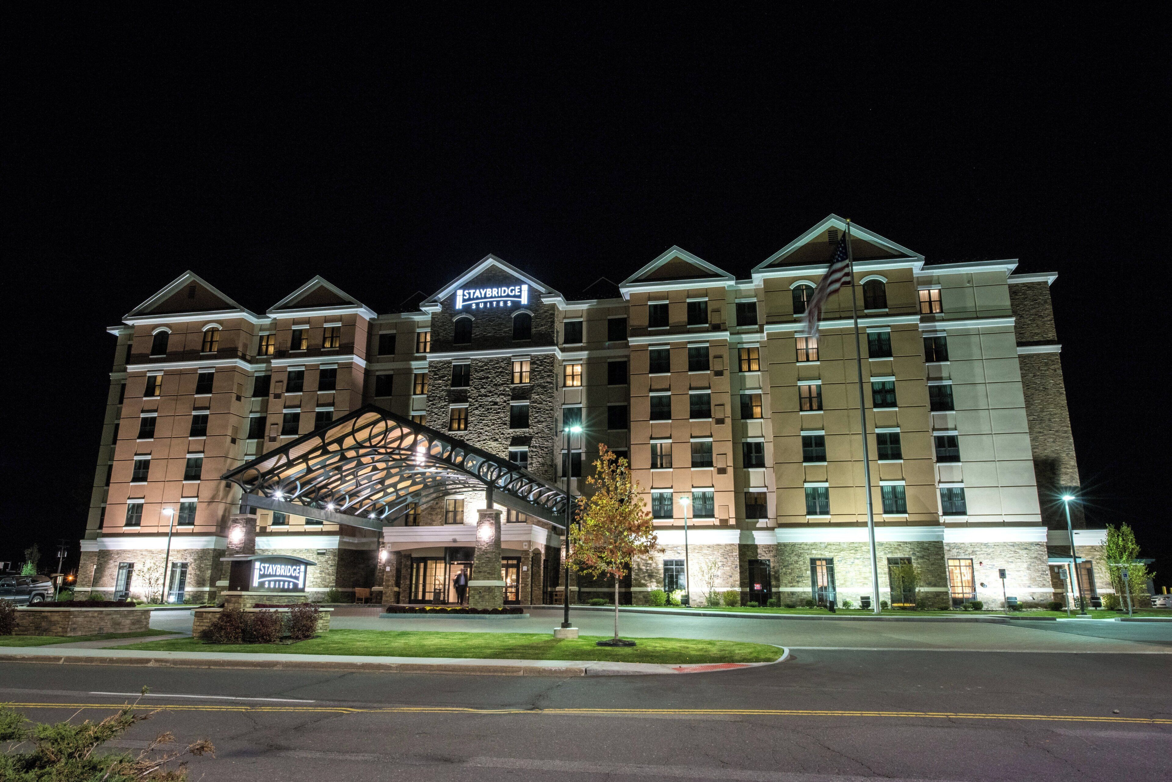 Un hotel de varios pisos con un llamativo letrero de Staybridge Suites, una entrada techada y una fachada bien iluminada.