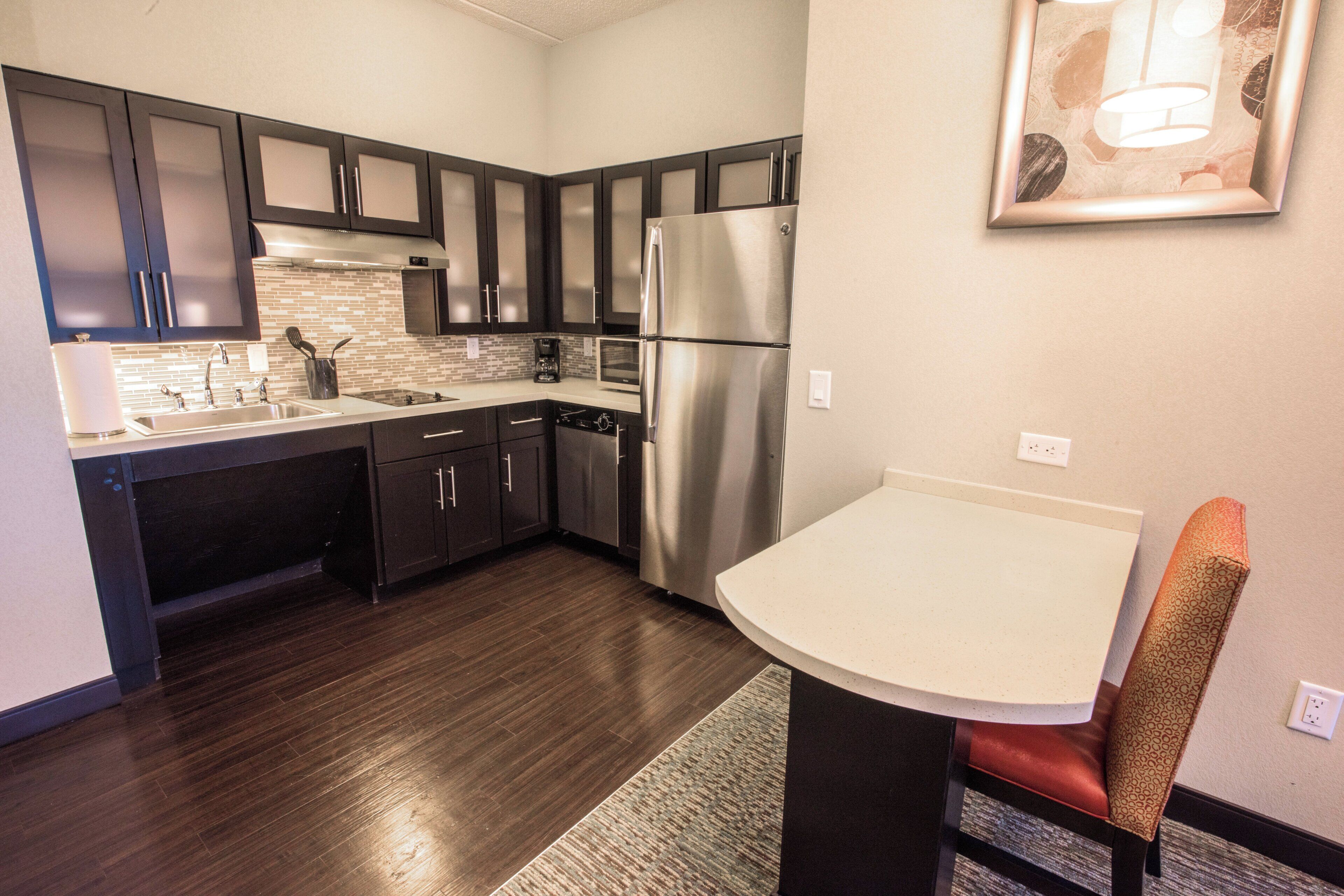 A modern kitchen with dark cabinetry, stainless steel appliances, and a small dining area with a white table and a patterned chair.