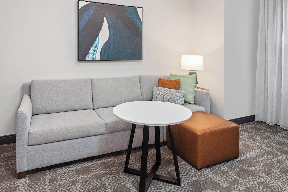 Une chambre d’hôtel moderne avec un canapé gris, une table ronde blanche et un pouf marron. Une lampe et des coussins colorés sont posés sur le canapé.