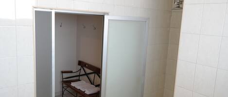 A small bathroom with white tiled walls, a wooden bench with a towel, and a shower area.