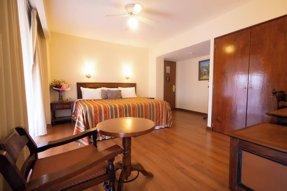 Hotelkamer met een bed, een bureau, een stoel, een ronde tafel, een plafondventilator en een nachtkastje met een bloevas.