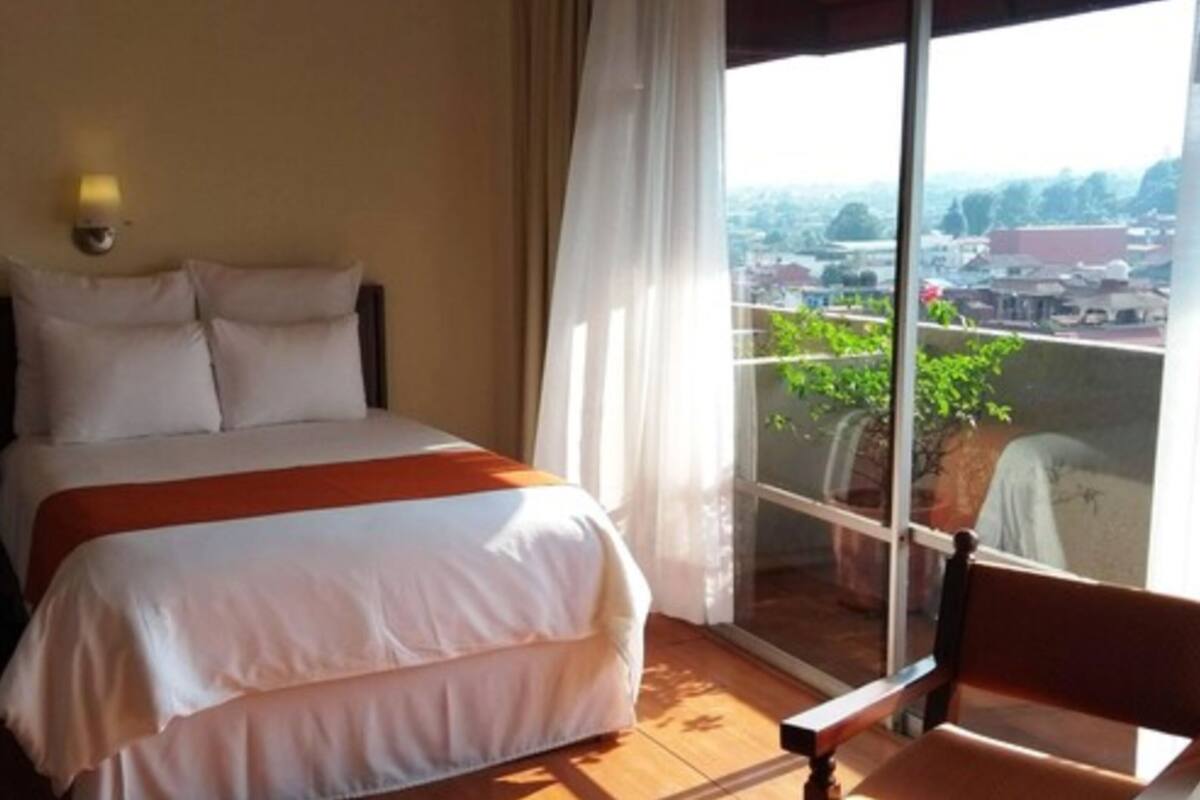 Een hotelkamer met een bed, een raam met uitzicht op een stadsbeeld en een balkon met potplanten.