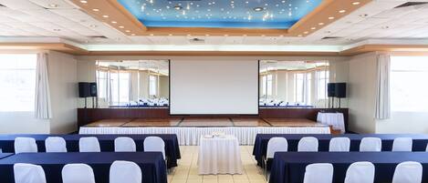 Una sala conferenze con un palco, file di tavoli con sedie, un grande schermo e un soffitto azzurro con luci.