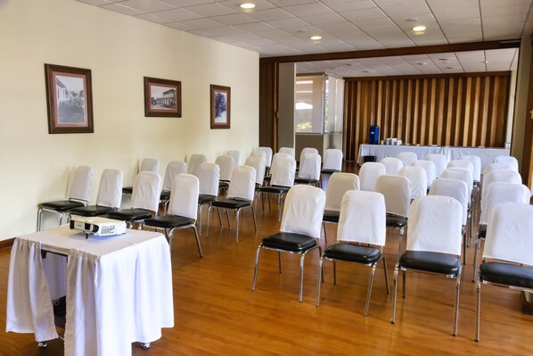 Un salón de conferencias con filas de sillas y mesas blancas, suelo de madera y cuadros en las paredes.