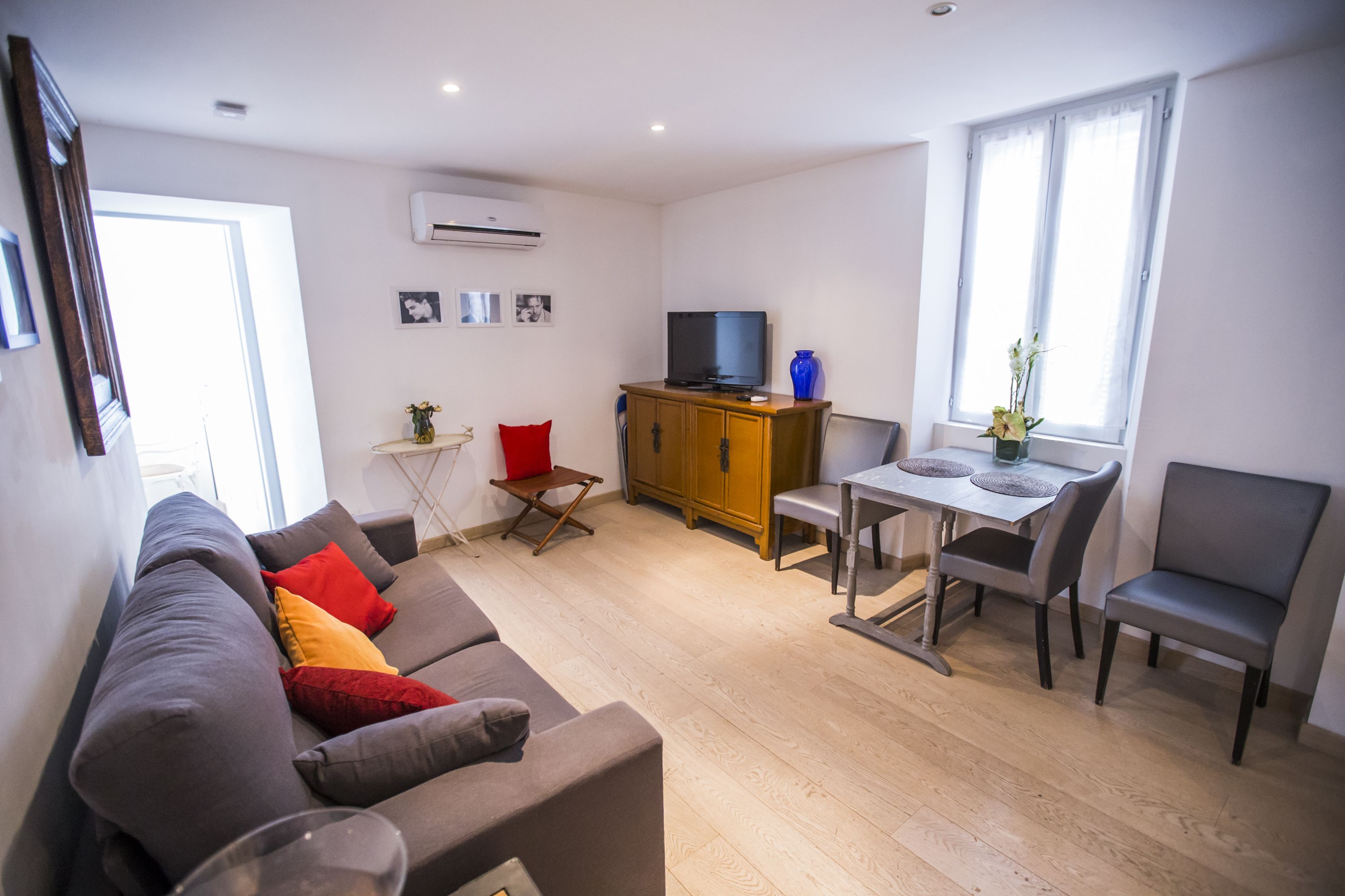 A modern living room with a grey sofa, a wooden cabinet, a dining table with chairs, and a TV.