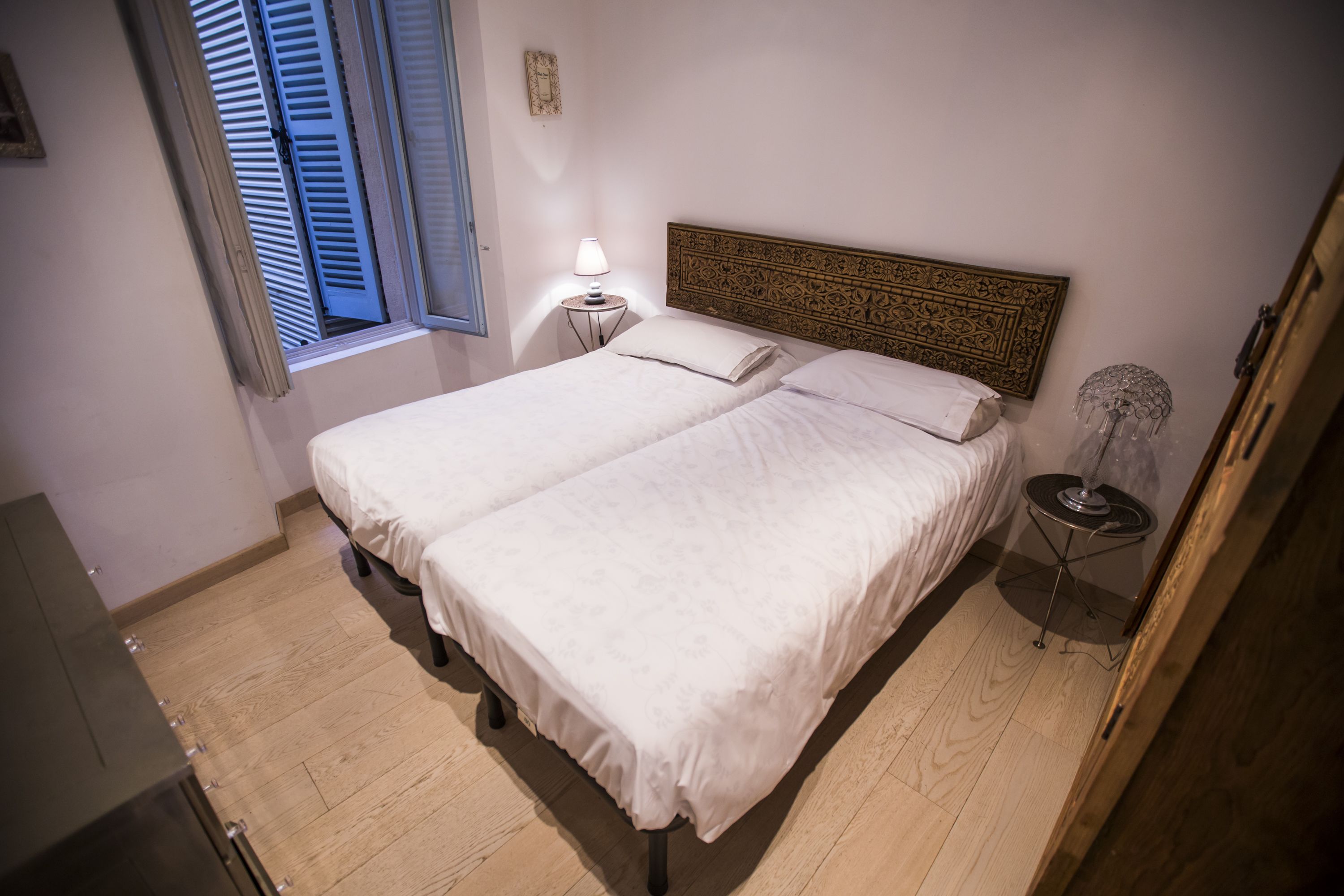 Una cama doble con cabecera, una mesita de noche con lámpara y una ventana con contraventanas.
