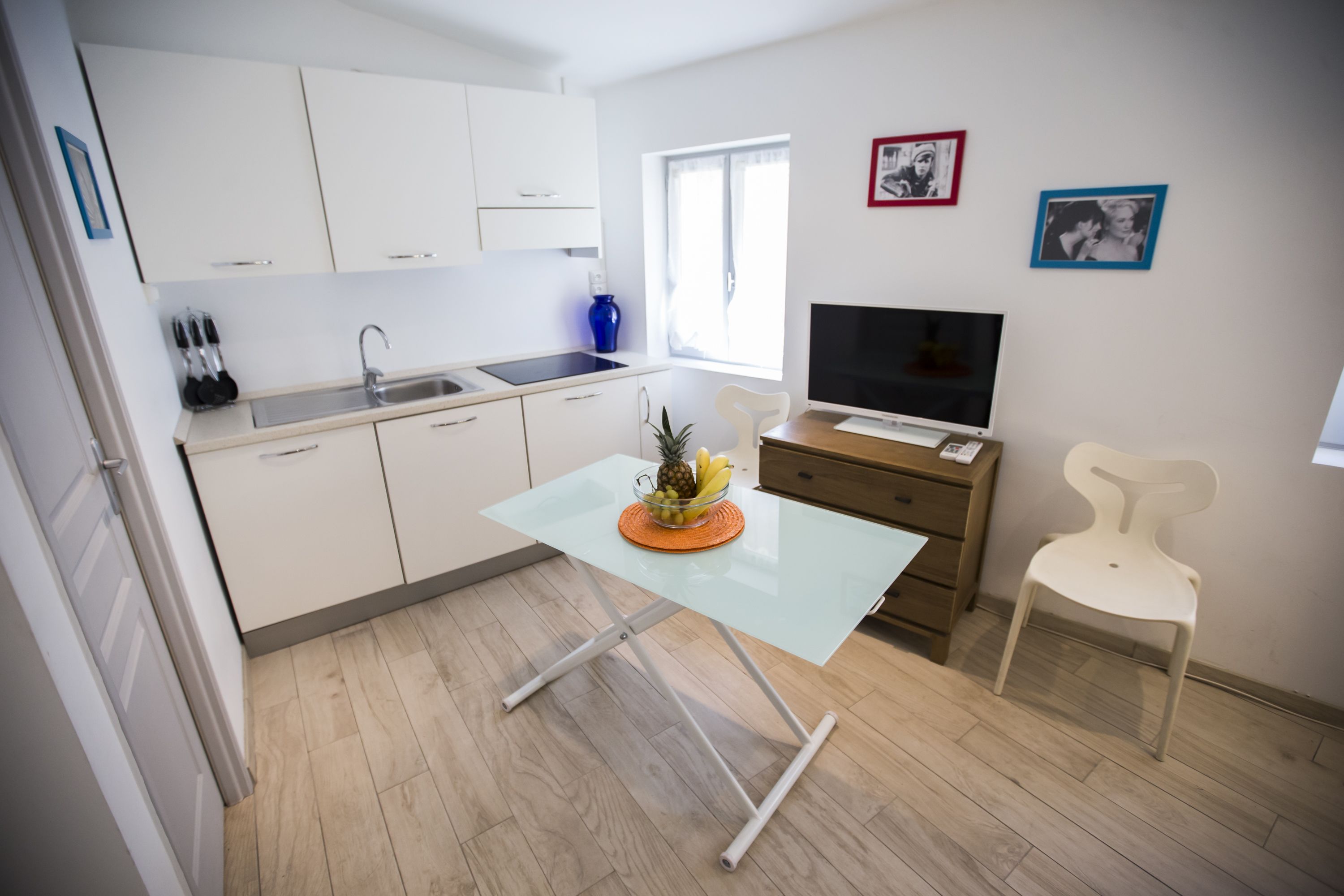 A modern kitchen with white cabinets, a glass table, a fruit bowl, a TV, and a chair.