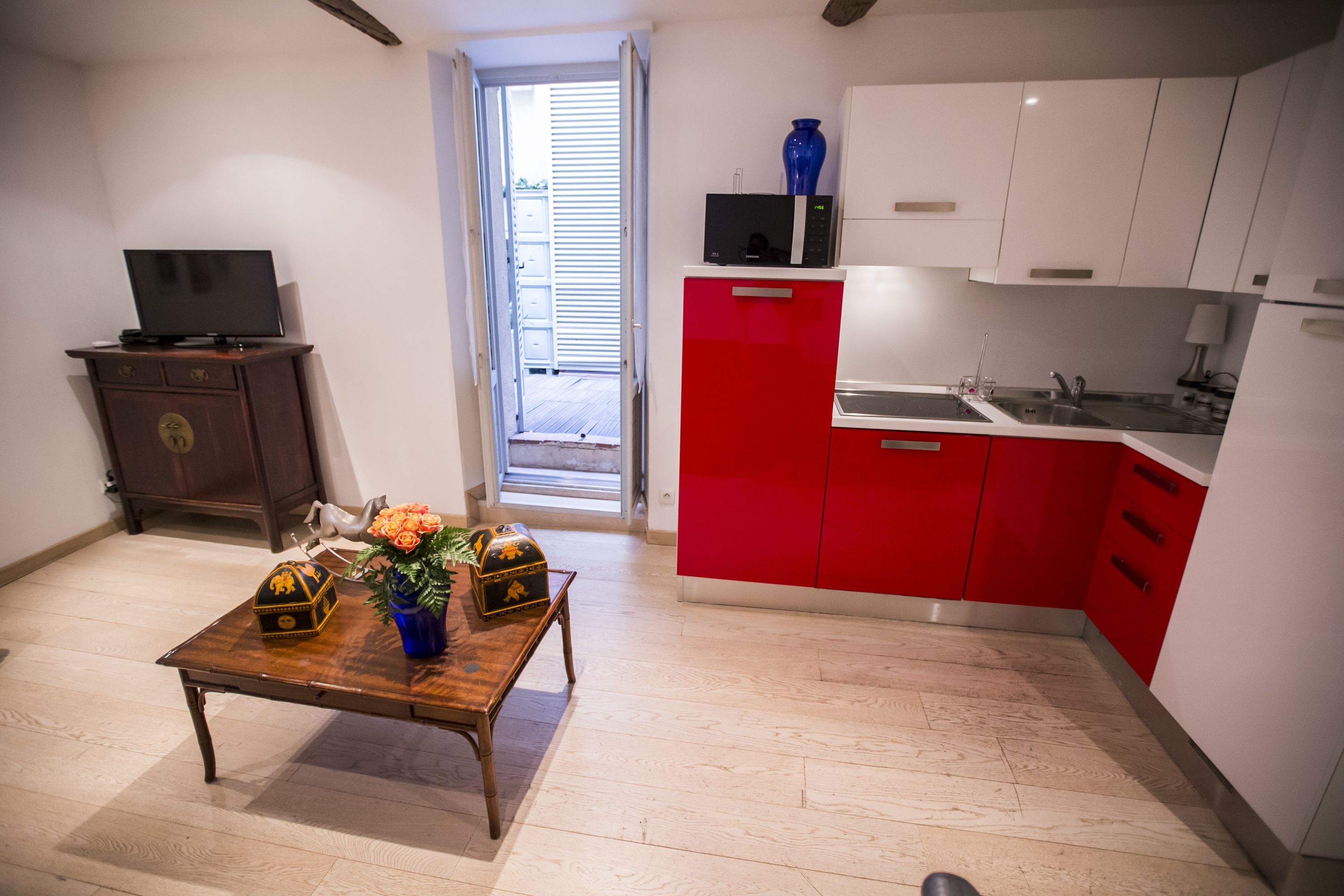 Une cuisine de taille réduite avec des meubles de rangement rouges et blancs, une table en bois ornée d’un vase et un téléviseur posé sur un meuble.
