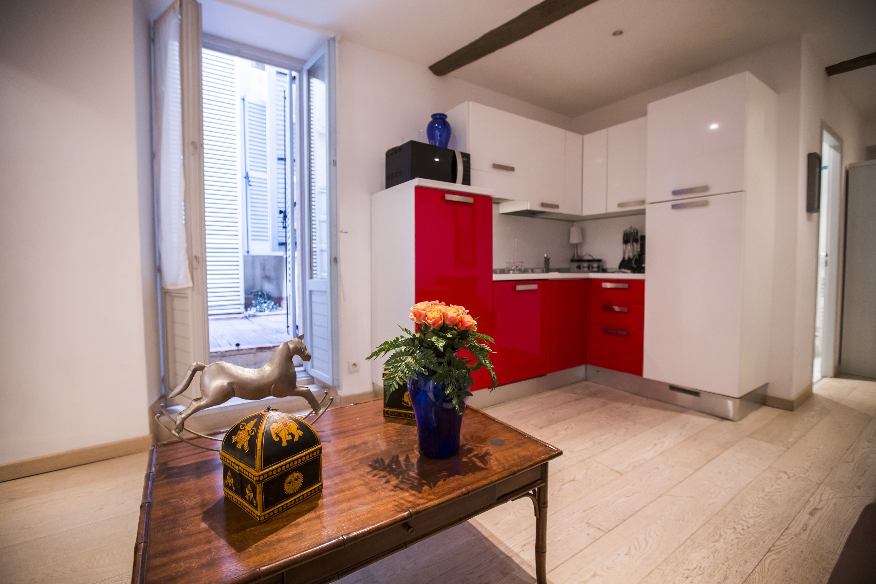 Een moderne keuken met witte en rode kasten, een houten eettafel met een decoratieve doos en vaas, en een raam met zonwering.