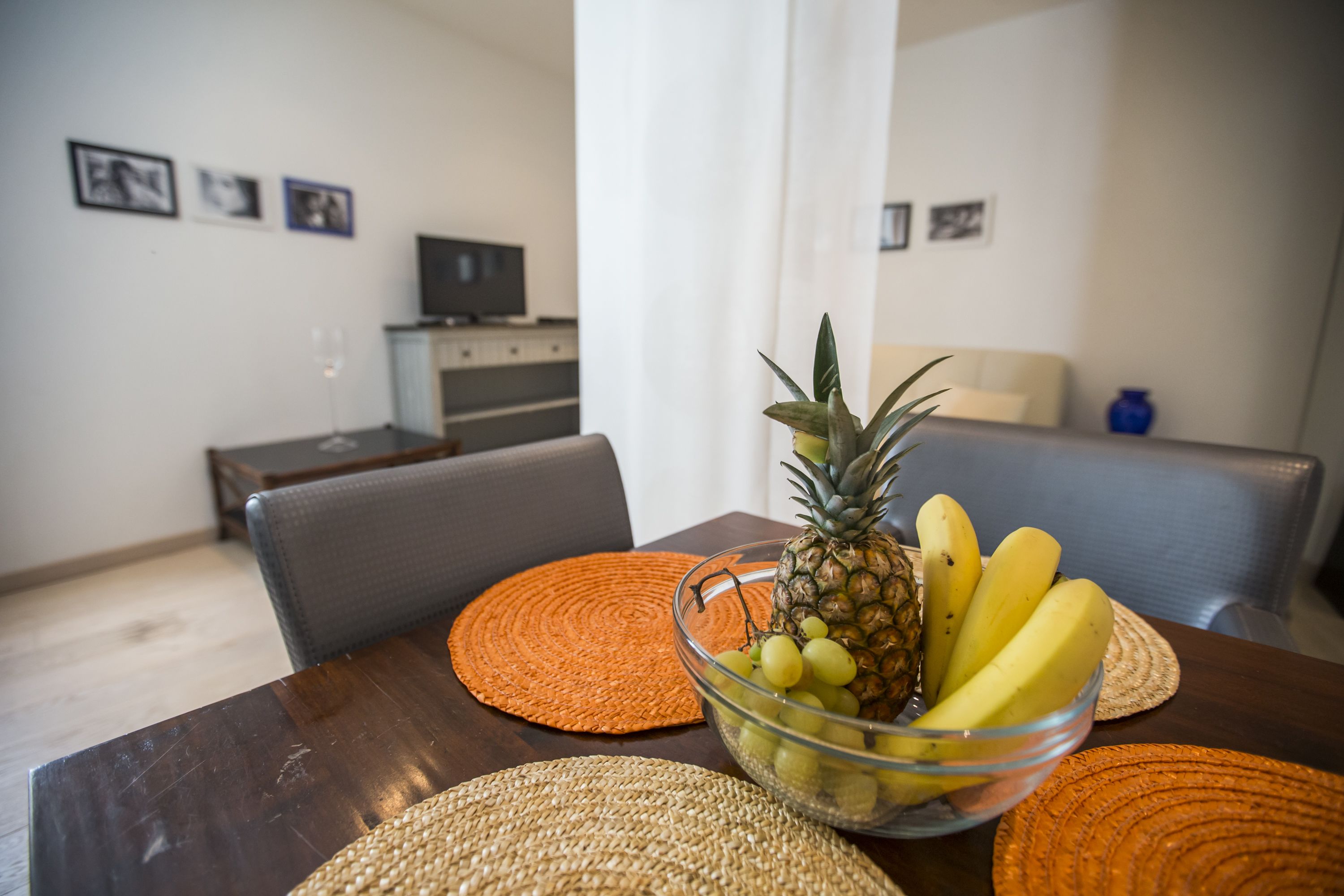 Ein Esstisch mit Obstschale, orangefarbenen Platzsets und einem Fernseher im Hintergrund.