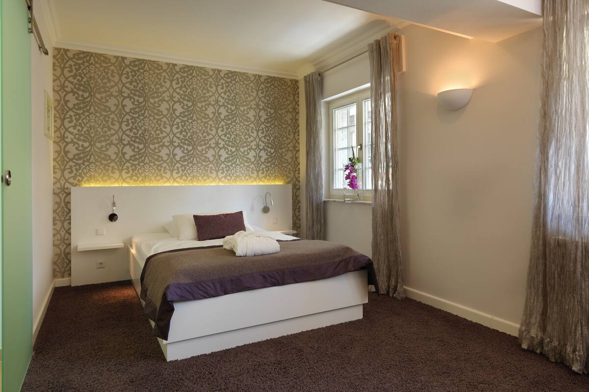 Une chambre moderne avec un grand lit, une table de chevet, une lampe fixée au mur et une fenêtre avec des rideaux.