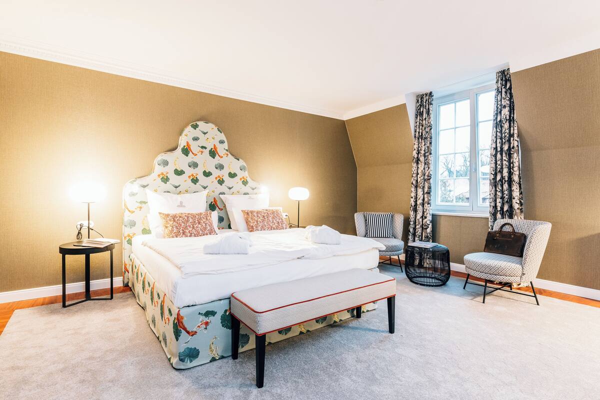 Une chambre avec un grand lit, une table de chevet, une chaise, une fenêtre avec des rideaux et une vue sur l’extérieur.