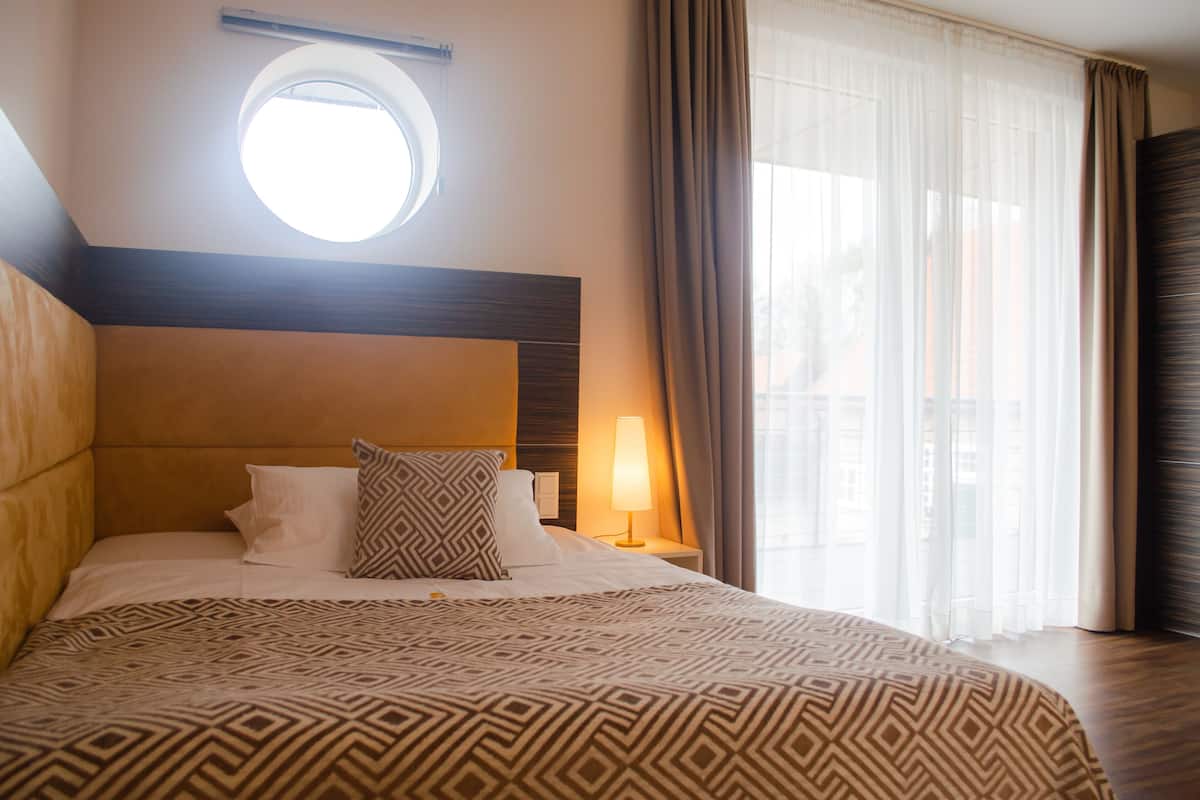 Une chambre d’hôtel avec un grand lit, une fenêtre ronde, une table de chevet avec une lampe et des rideaux légers.