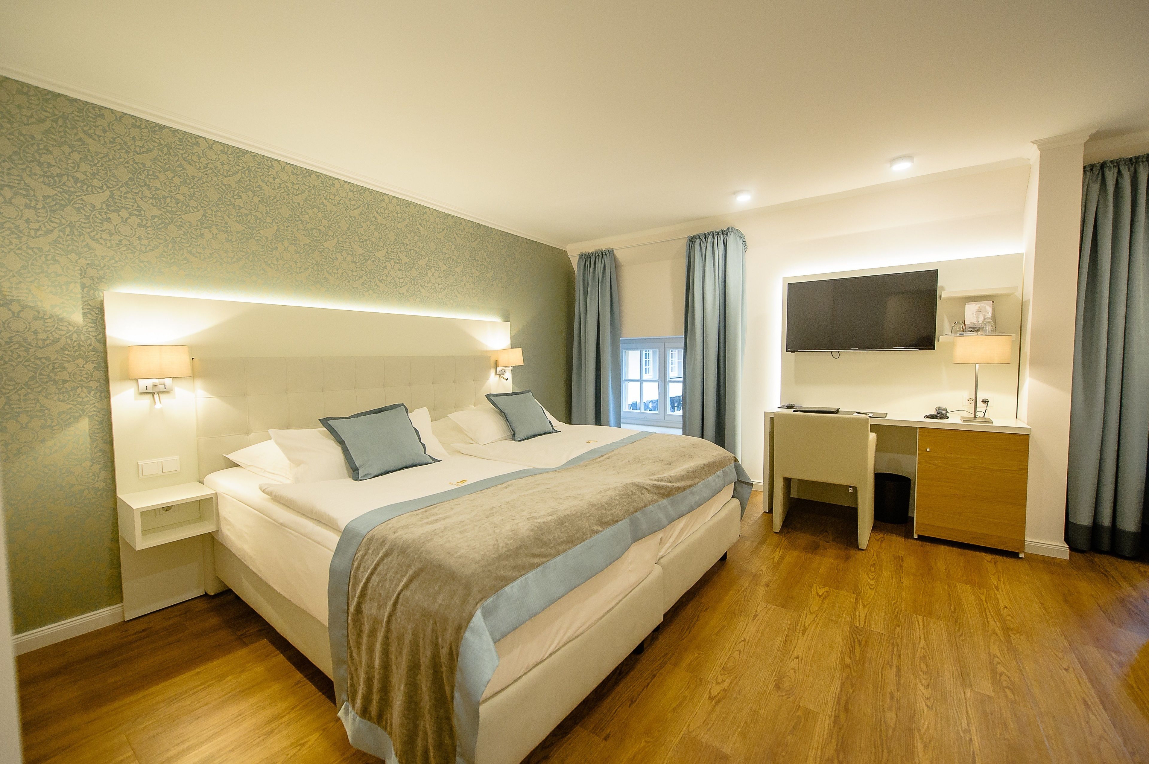 A hotel room with a large bed, bedside tables, a desk, a TV, and a window with curtains.