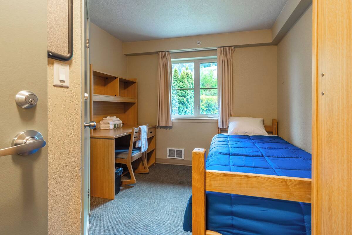A bedroom with a single bed, a desk with a chair, a wardrobe, and a window with curtains.