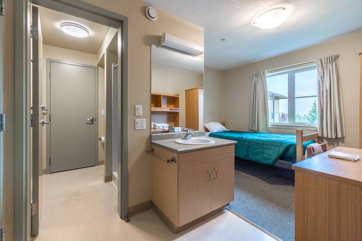 A hotel room with a bathroom, a bed with teal bedding, a wooden desk, and a window with curtains.