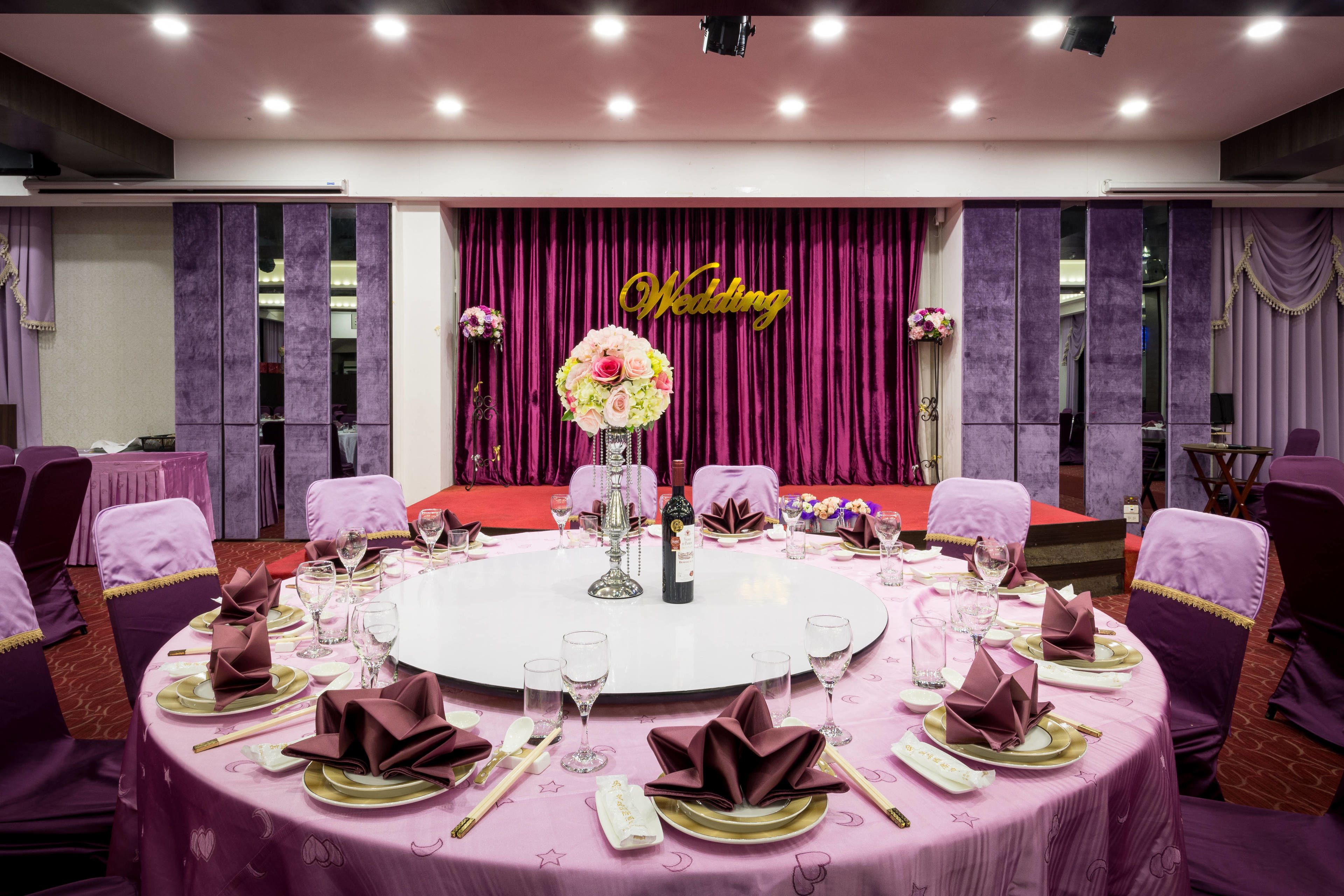 A round table set for a wedding reception with purple tablecloths, gold and purple napkins, and a floral centerpiece.