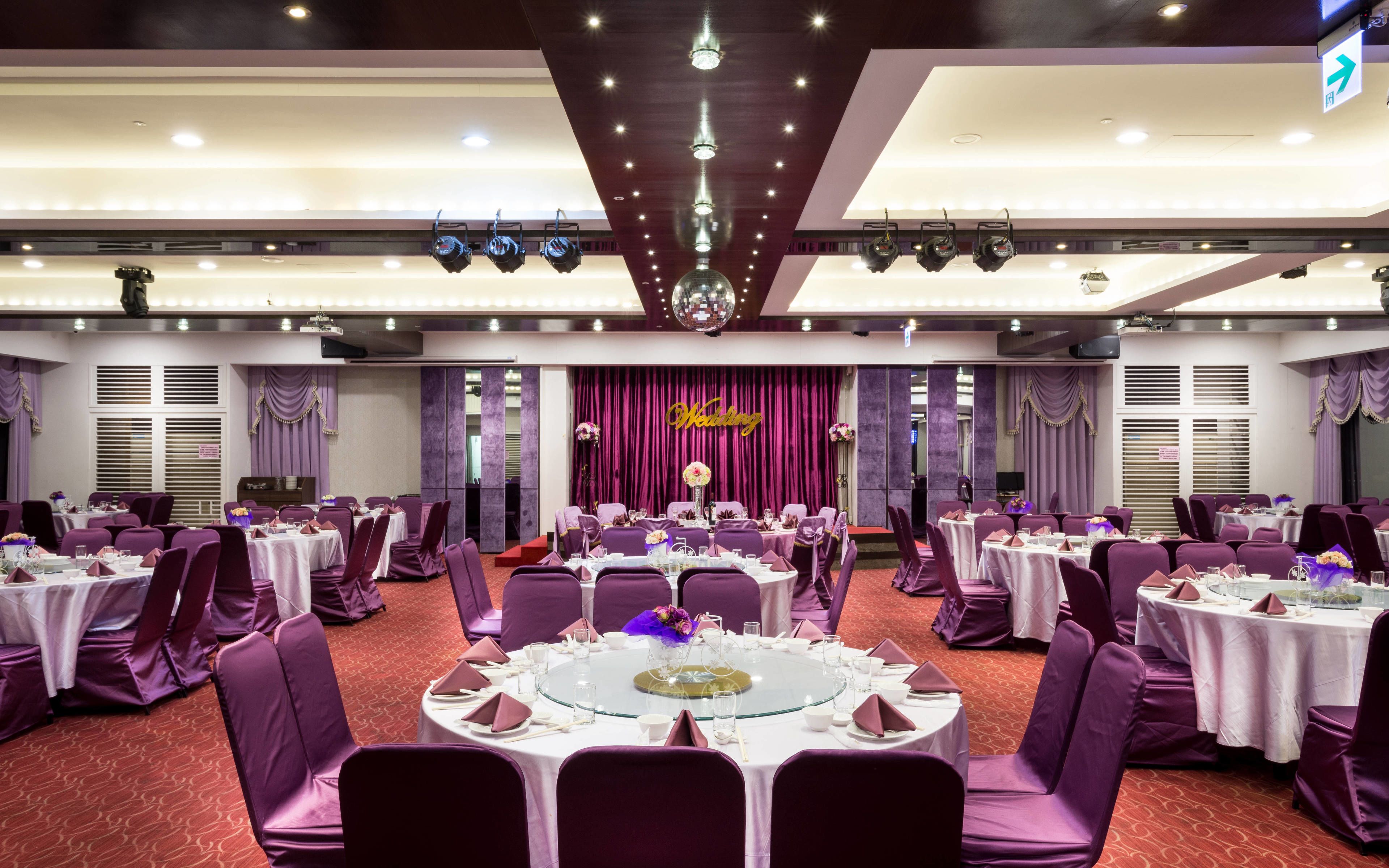 Un salón de banquetes con mesas redondas, sillas moradas y un escenario con un cartel que dice 'Wedding'.